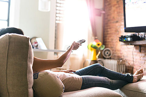 Frau sitzt zurückgelehnt auf einem Sofa vor dem Fernseher und hat eine Fernbedienung in der Hand