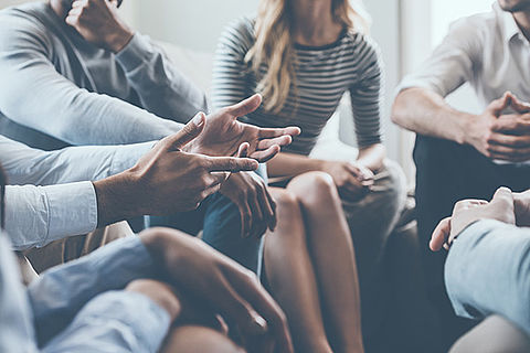 Mehrere Leute sitzen im Kreis, es sind allerdings hauptsächlich nur die Hände zu sehen. Sie gestikulieren stark, also wird vermutlich diskutiert.