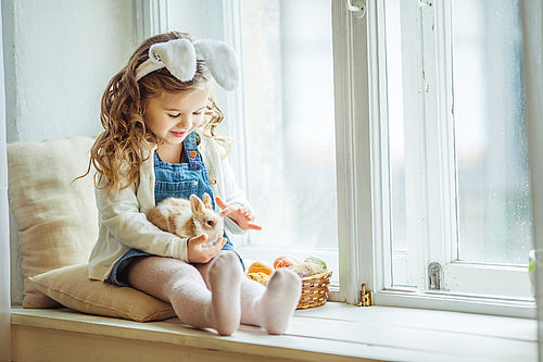 Ein kleines Mädchen sitzt auf einer Fensterbank. Sie hat ein Kaninchen auf dem Schoß und Osterhasenohren auf dem Kopf. Frohe Ostern!