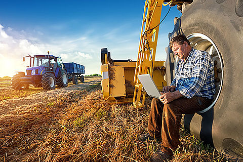 Ein Landwirt sitzt in einem Traktorreifen. Weitere Erntemaschinen sind im Hintergrund zu sehen. Der Landwirt schaut auf einen Laptop.