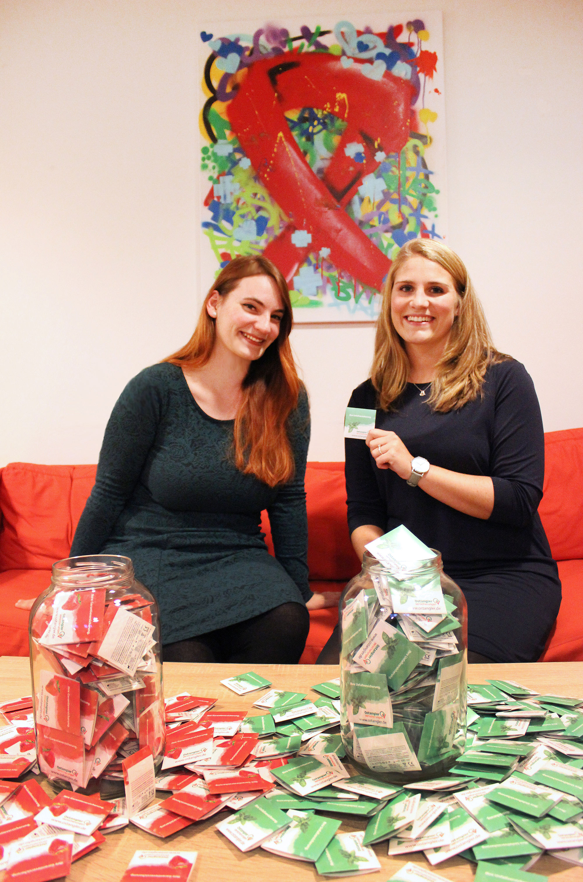 Frau Wollenberg (Ostangler) über eine Spende an die Flensburger AIDS-Hilfe. Auf dem Tisch vor den beiden Frauen sind haufenweise grüne und rote Kondome ausgebreitet, die Farben des Ostangler-Logos.