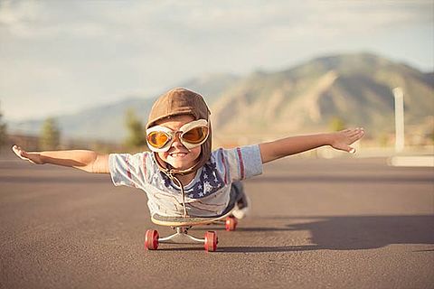 ein kleiner Junge fährt Skateboard, allerdings liegt er dafür flach auf dem Bauch, die Arme zu beiden Seiten ausgestreckt. Er trägt außerdem eine Fliegerbrille und einen Fliegerhelm.