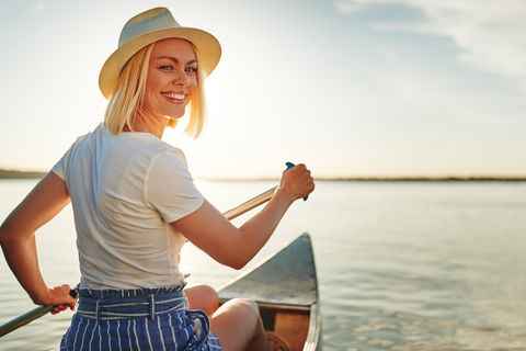 Blonde Frau mit Strohhut rudert übers Wasser und dreht sich leicht um, um den Betrachter anzulächeln. Hinter ihr strahlt die Sonne überm Horizont.