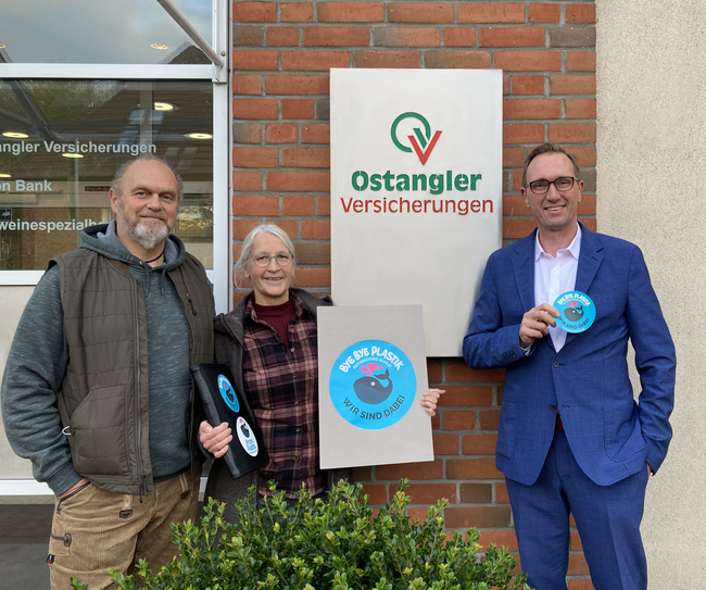 Herr Rohwer (Vorstandsvorsitzender Ostangler) hält das Abzeichen "Bye bye Plastik - wir sind dabei!" hoch. Neben ihm stehen zwei Vertreter der Organisation. Die 3 stehen vor dem Ostangler Bürogebäude und lächeln in die Kamera.