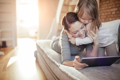 Eine Mutter und ihre junge Tochter entspannen sich zusammen auf dem Sofa. Beide schauen zusammen etwas auf dem Tablet.
