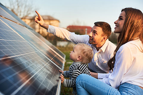 junger Familie (Vater, Mutter, Kleinkind) hocken vor Solarpanel einer Photovoltaikanlage, um sie sich näher anzusehen