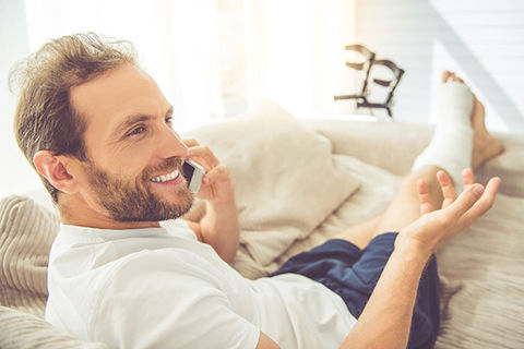 Ein Mann liegt auf der Couch mit einem eingegipsten Bein. Er telefoniert und macht ein fröhliches, sorgenfreies Gesicht.