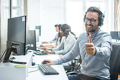 Ein Mitarbeiter sitzt vor seinem Computer mit einem Headset auf dem Kopf. Er dreht sich zum Betrachtet, lächelt ihn and und gibt ihm sogar ein "Daumen hoch!". Im Hintergrund sind weitere Mitarbeiter*innen zu sehen.