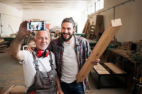 Ein älterer und ein jüngerer Mann machen ein Selfie. Sie stehen inmitten einer Werkstatt, der junge Mann hält ein Stück Holz hoch. Vermutlich sind sie Schreiner.