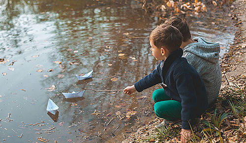 Zwei Jungen hocken vor einer großen Pfütze, vor ihnen treiben zwei Papierboote. Einer der Jungen stupst sein Boot mit einem Stock an.