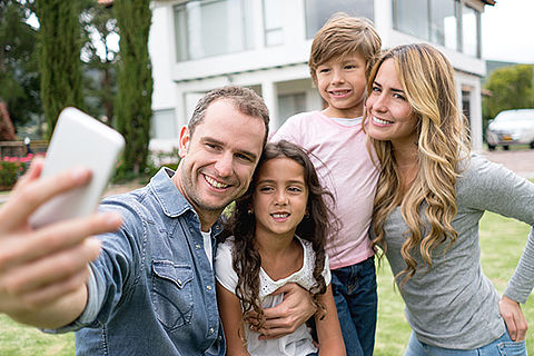 Vater, Mutter, Tochter und Sohn machen ein Familienfoto mit dem Handy vor ihrem vermutlich neuen Haus.