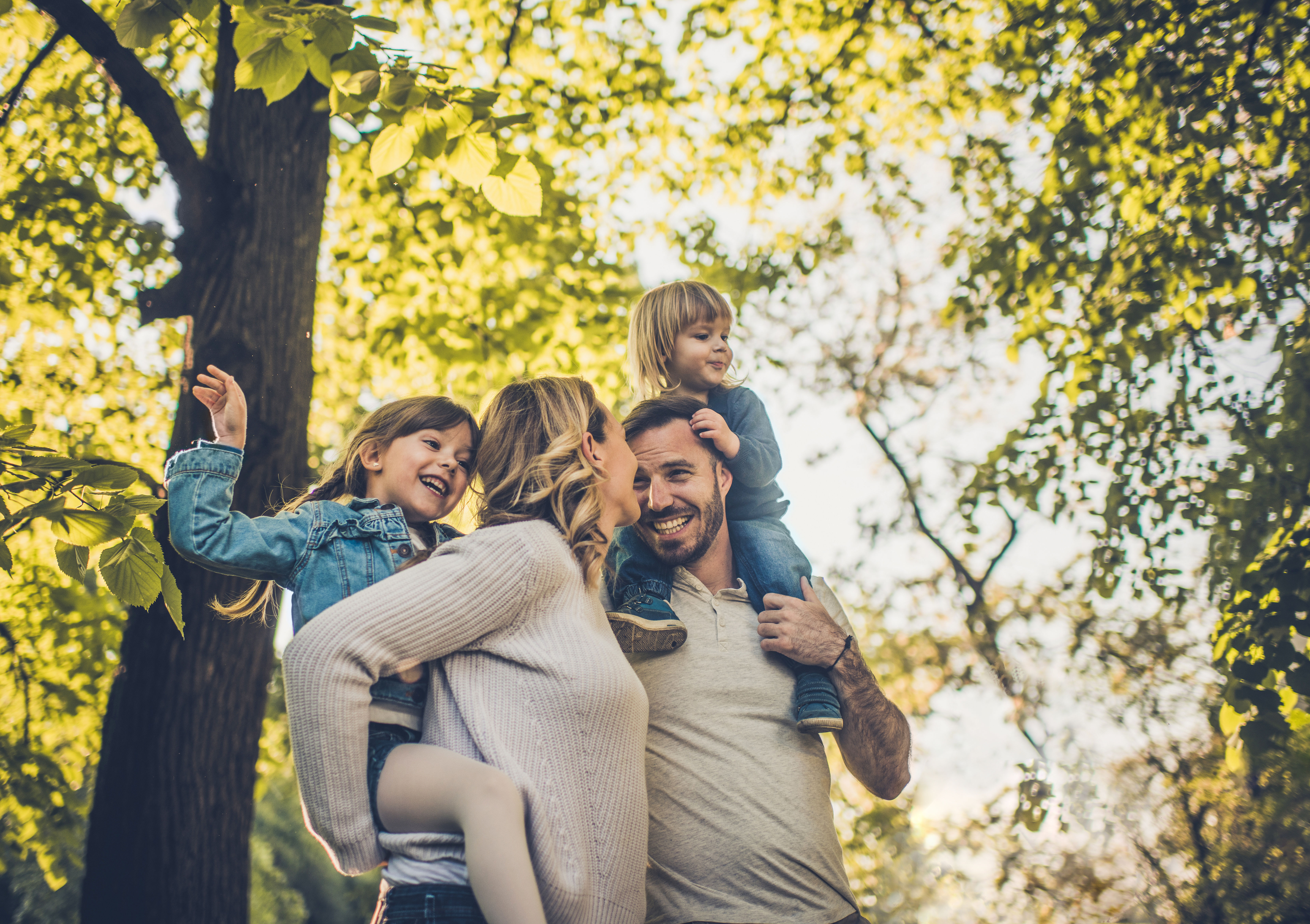 Junge Familie macht einen Waldspaziergang, der Vater hat ein Kleinkind auf den Schultern, die Mutter trägt das zweite Kind Huckepack.