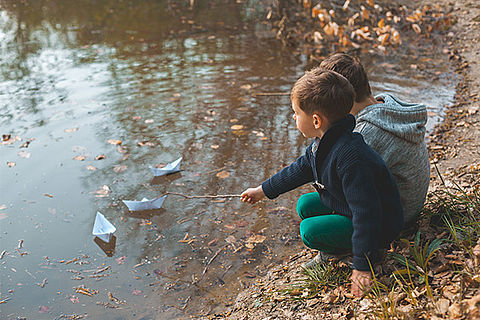 Zwei Jungen hocken vor einer großen Pfütze, vor ihnen treiben zwei Papierboote. Einer der Jungen stupst sein Boot mit einem Stock an.