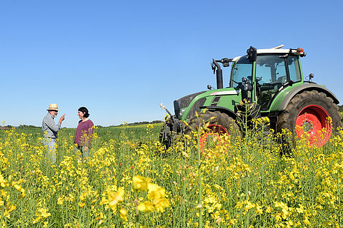 Zwei Landwirte streiten sich. Sie stehen inmitten eines blühenden Rapsfeldes, ein grüner Traktor steht neben ihnen.