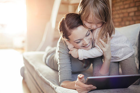 Mutter und Tochter liegen entspannt auf dem Sofa und schauen dabei gemeinsam etwas auf dem Tablet an.